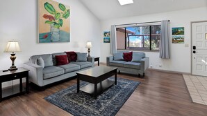 A living room with two grey sofas, a coffee table, and a rug. There are two lamps, a painting on the wall, and a window with curtains. - La Selva Beach Retreat Near Manresa Beach (Watsonville)
