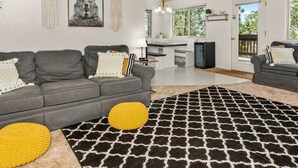 A modern living room with a grey sofa, yellow pouf, and a black and white patterned rug. There is a kitchen area with white cabinets and a black refrigerator. - Shaver Lake Cabin with Deck and Mountain Views (Auberry)