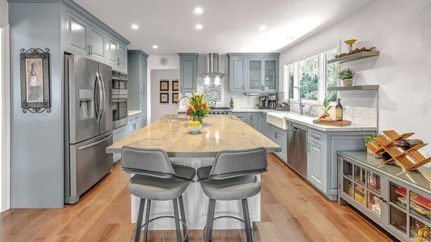 A modern kitchen with grey cabinets, stainless steel appliances, a large island with bar stools, and a window overlooking greenery.