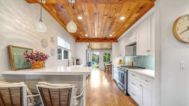 A modern kitchen with white cabinetry, a wooden ceiling, and a central island with chairs. - Del Monte Beach Cottage Sleeps 4 with Private Yard (Seaside)