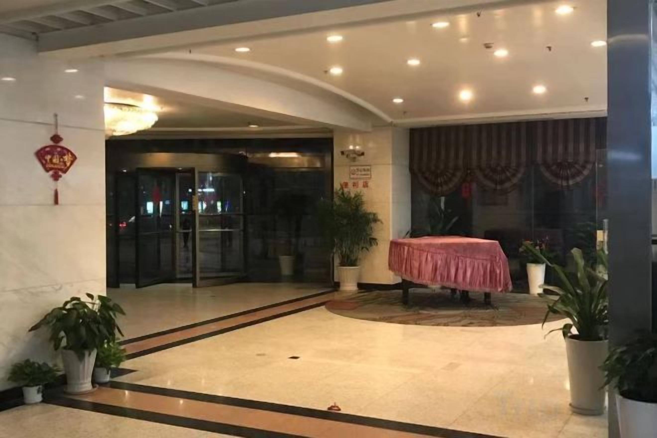 Um hall de hotel com balcão de recepção, entrada por porta de vidro, plantas em vasos e uma área de refeições com mesa e cadeiras.