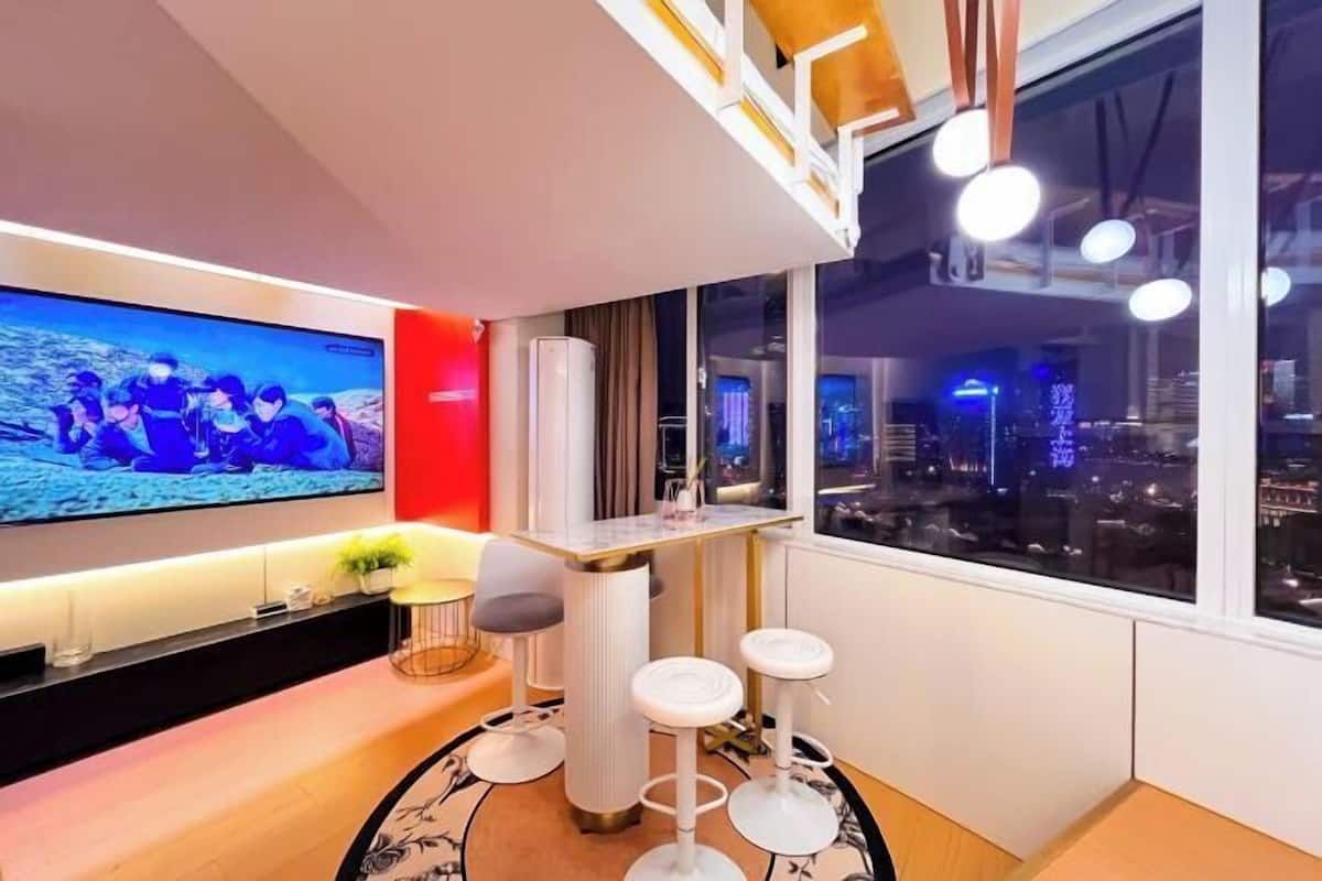 A modern hotel room with a large TV, a dining area with stools, and a city view through large windows.