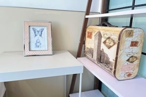Une œuvre d’art représentant un papillon, encadrée, posée sur une table blanche, une valise ancienne ornée de tampons de voyage et une fenêtre avec des stores.
