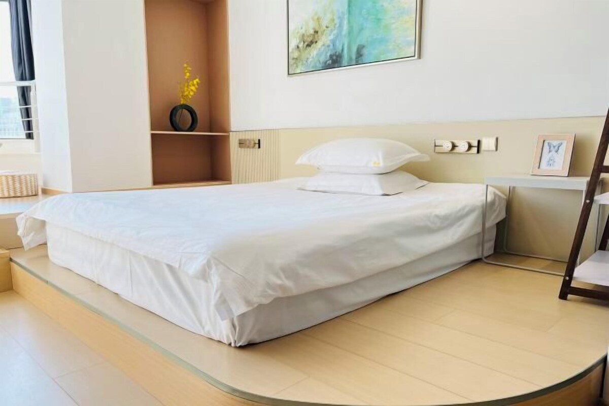 Un lit bien fait dans une chambre d’hôtel moderne, avec un plancher incurvé, une petite étagère ornée d’un vase de fleurs et un tableau accroché au mur.