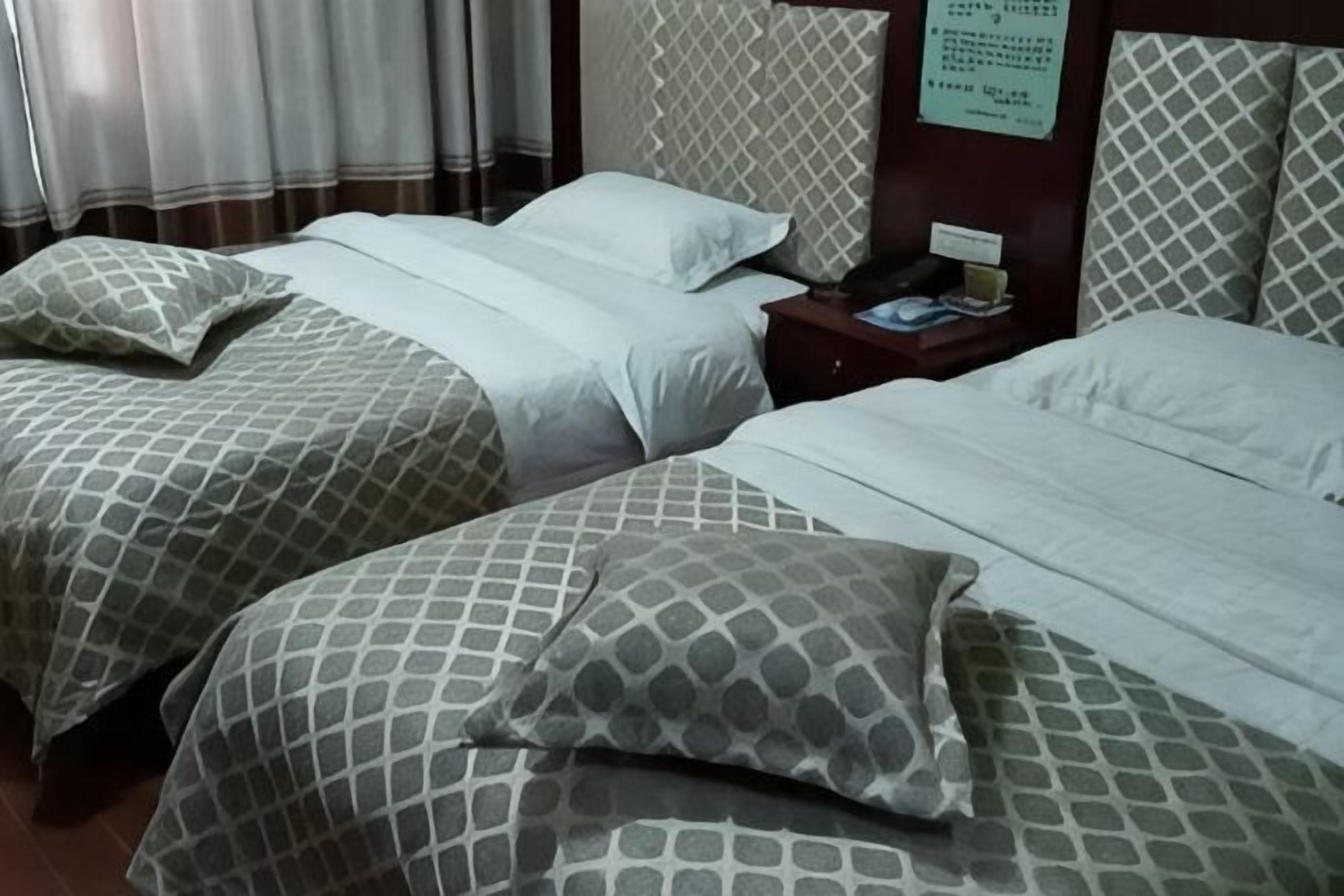 Duas camas com roupa de cama estampada, uma mesinha de cabeceira com abajur e um letreiro fixado na parede.