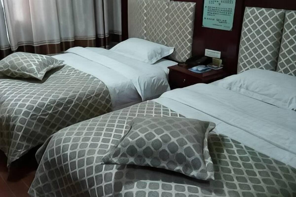 Duas camas com roupa de cama estampada, uma mesinha de cabeceira com abajur e um letreiro fixado na parede.