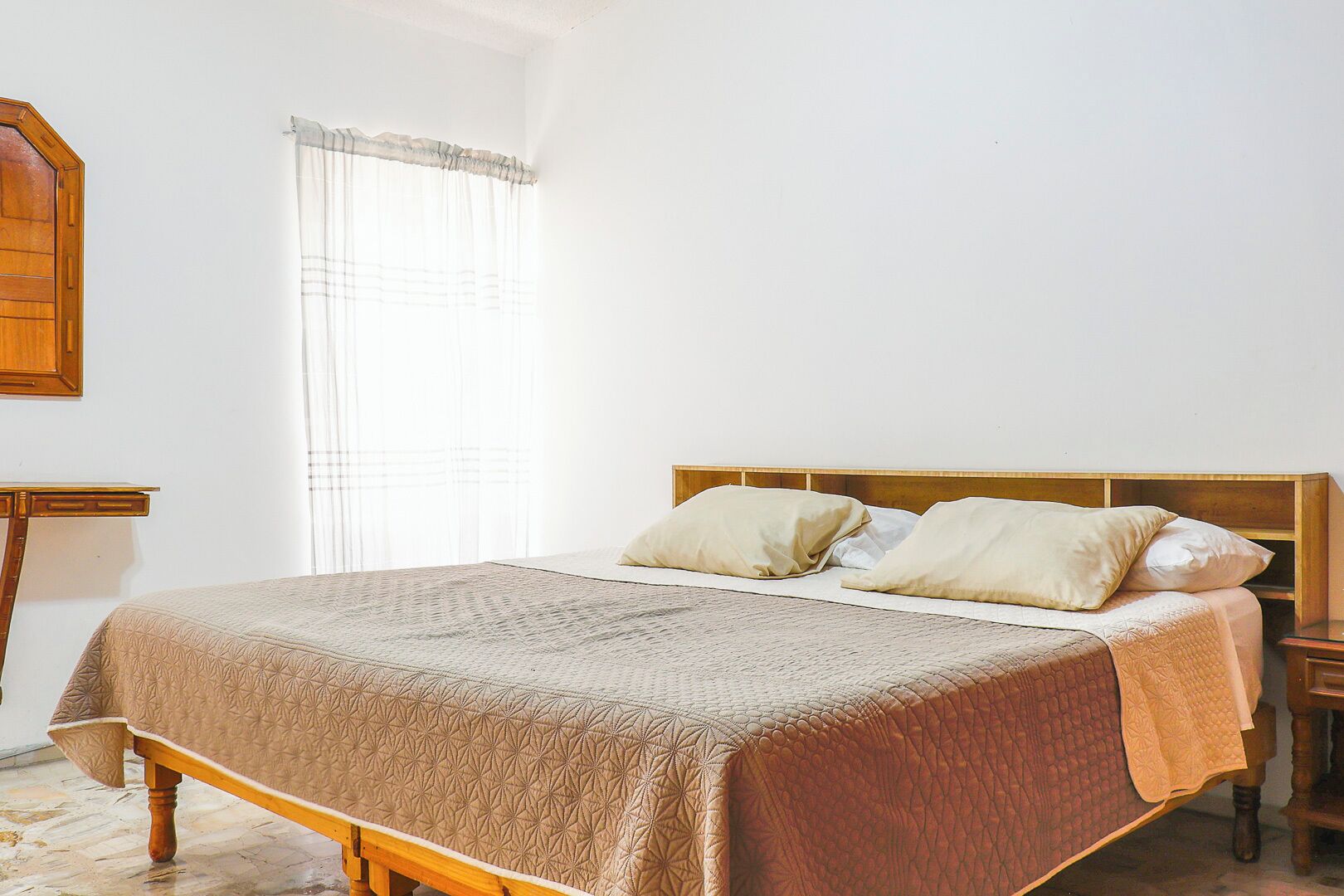 A bedroom with a wooden bed, two pillows, a bedside table, and a window with sheer curtains.
