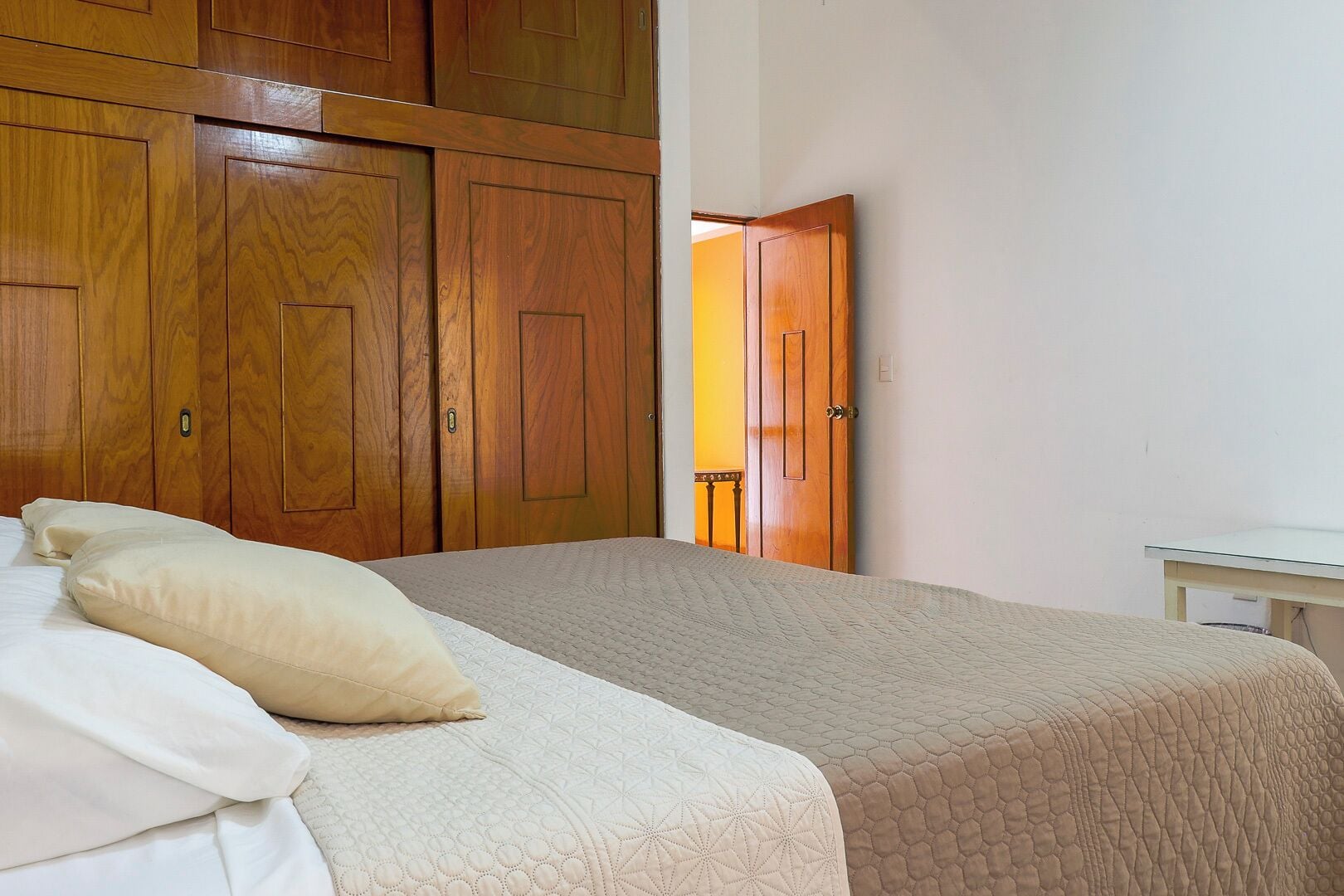 A bedroom with a bed, two pillows, a wooden wardrobe, and a small table.