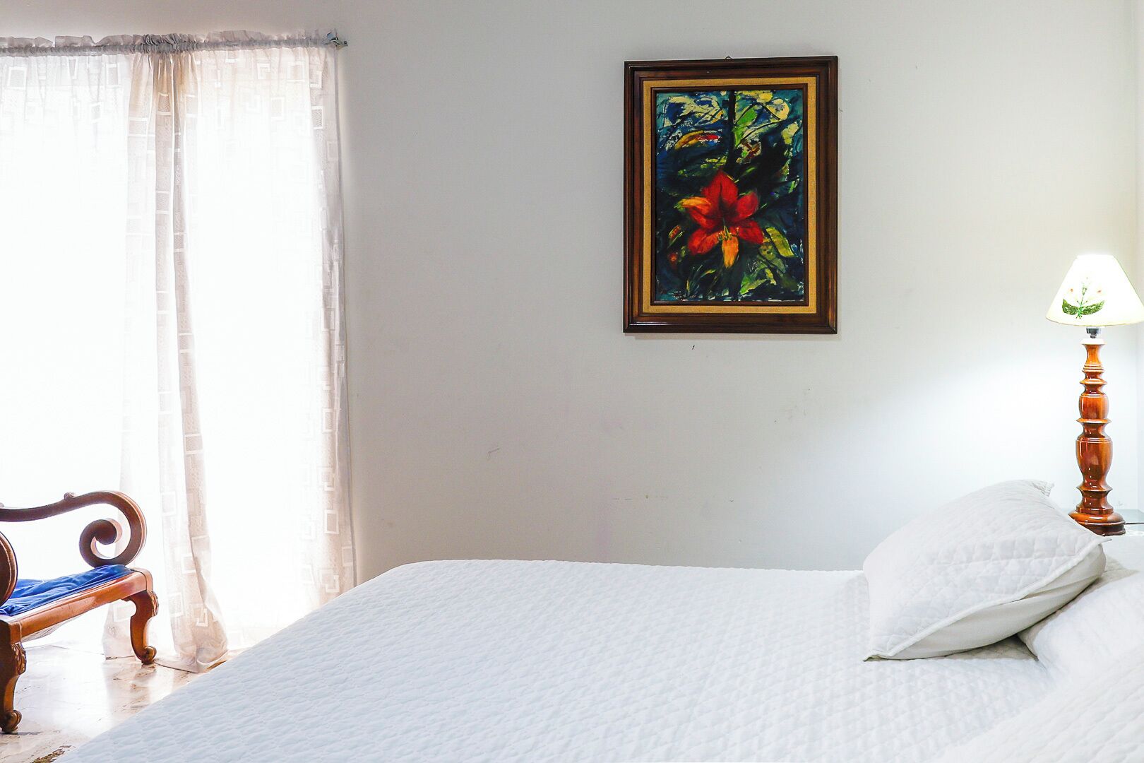 A neatly made bed with white bedding, a wooden chair, a table lamp, and a framed painting on the wall.