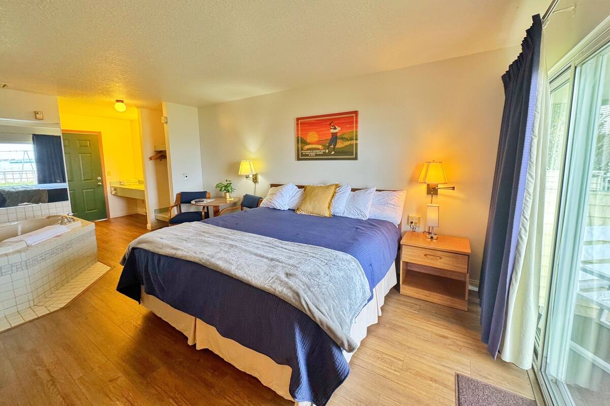 Habitación de hotel con una cama grande, mesitas de noche con lámparas, una pequeña mesa con una silla, un espejo y una puerta que da a otra habitación.