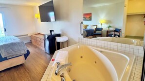 Une chambre d’hôtel avec une grande baignoire, un lit, un bureau avec une chaise, une petite table ronde, un miroir et un téléviseur fixé au mur.
