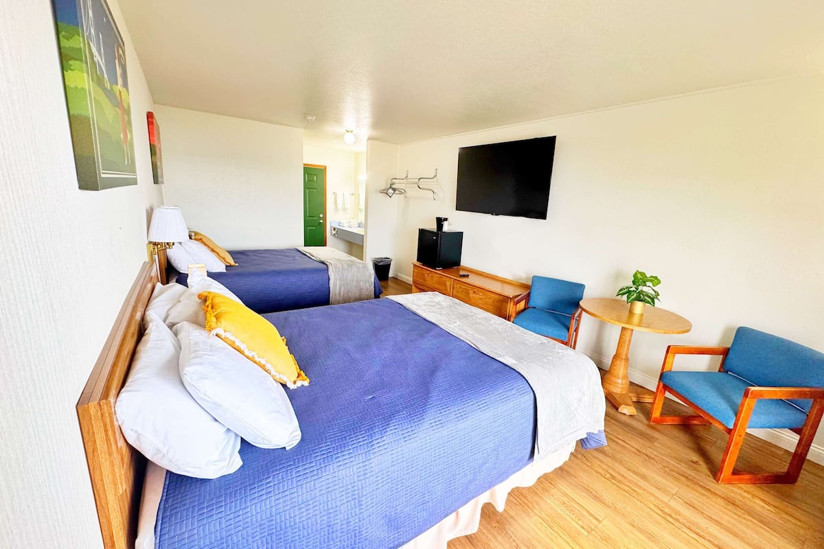 Habitación de hotel con dos camas, un televisor, una mesita con una silla y suelo de madera.