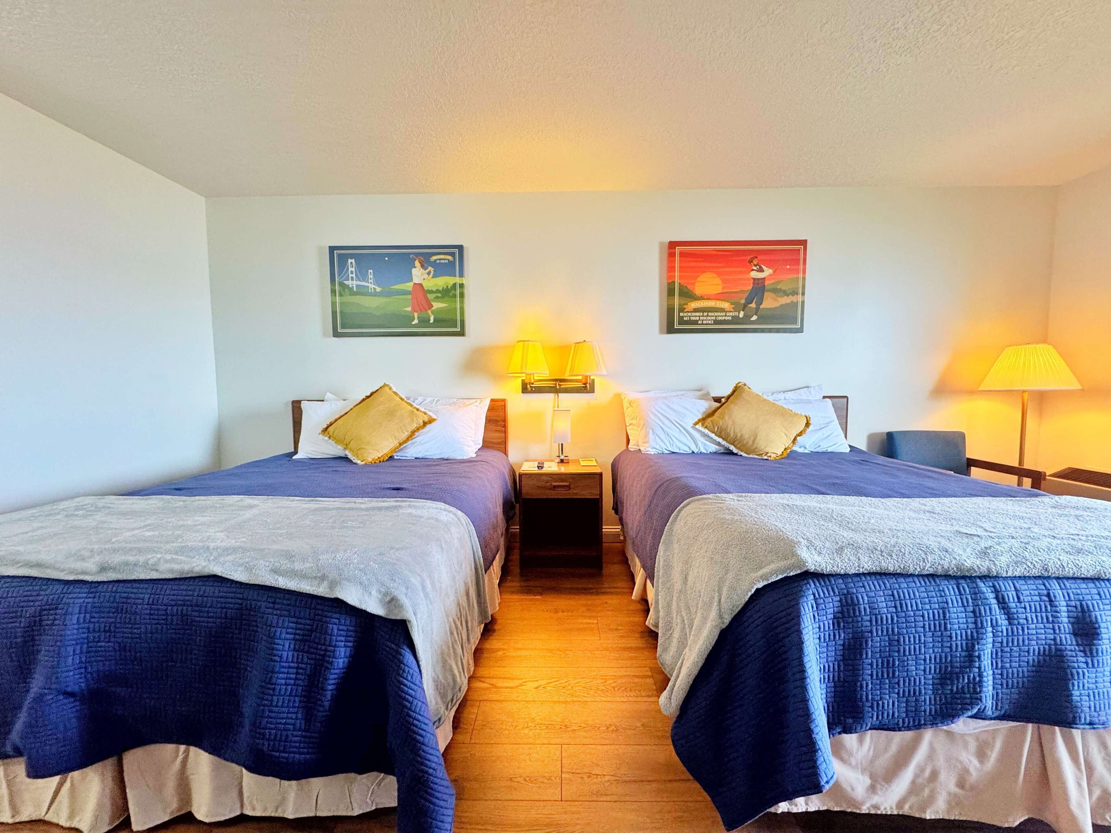 A hotel room with two beds, each with a blue bedspread and white pillows. There are two wall-mounted lamps on either side of the beds.