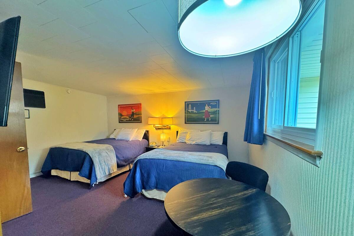 Una habitación de hotel con dos camas, una mesa redonda y un ventanal con cortinas.