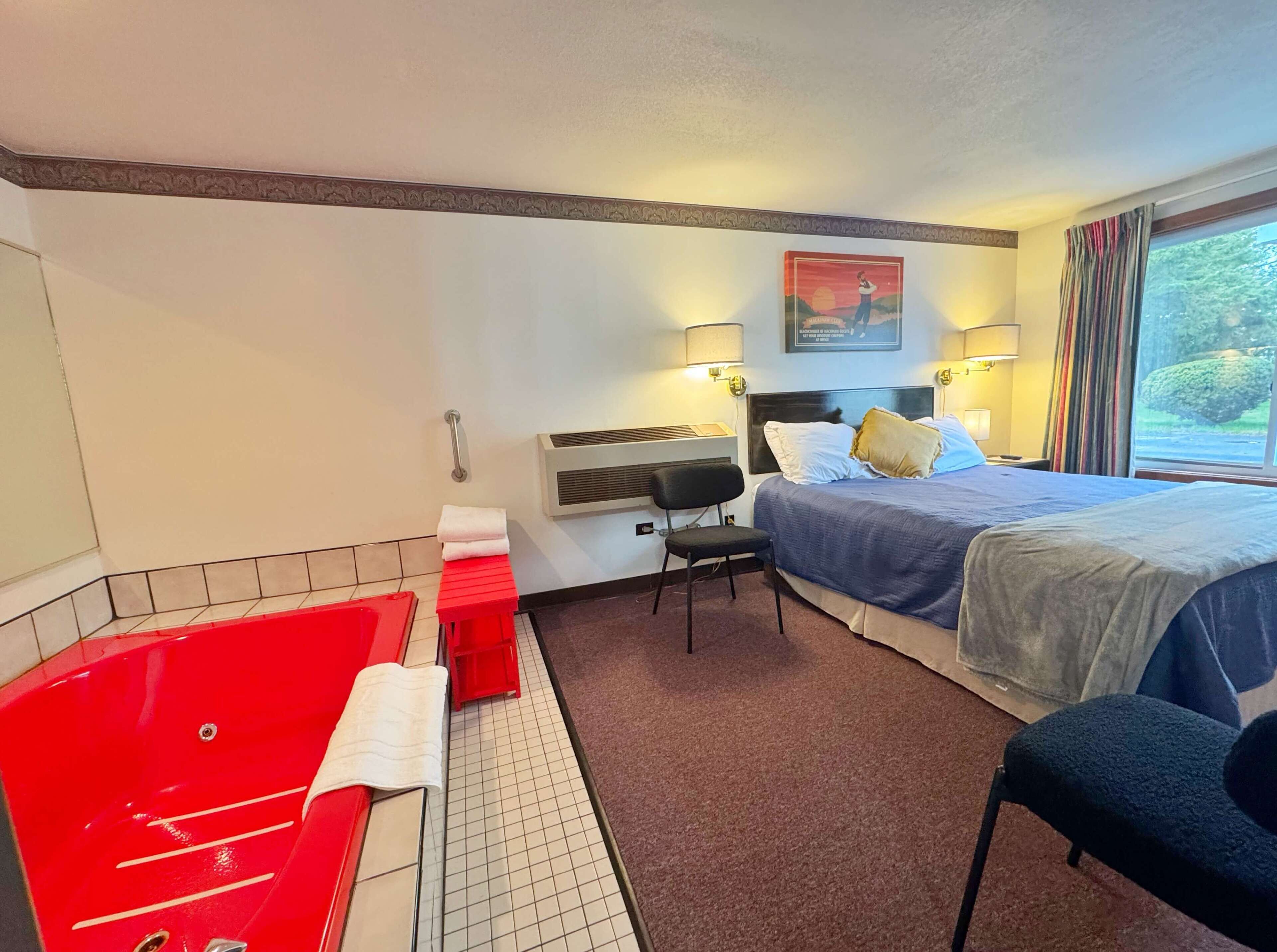 A hotel room with a large red bathtub, a bed with a blue bedspread, a chair, and a desk.