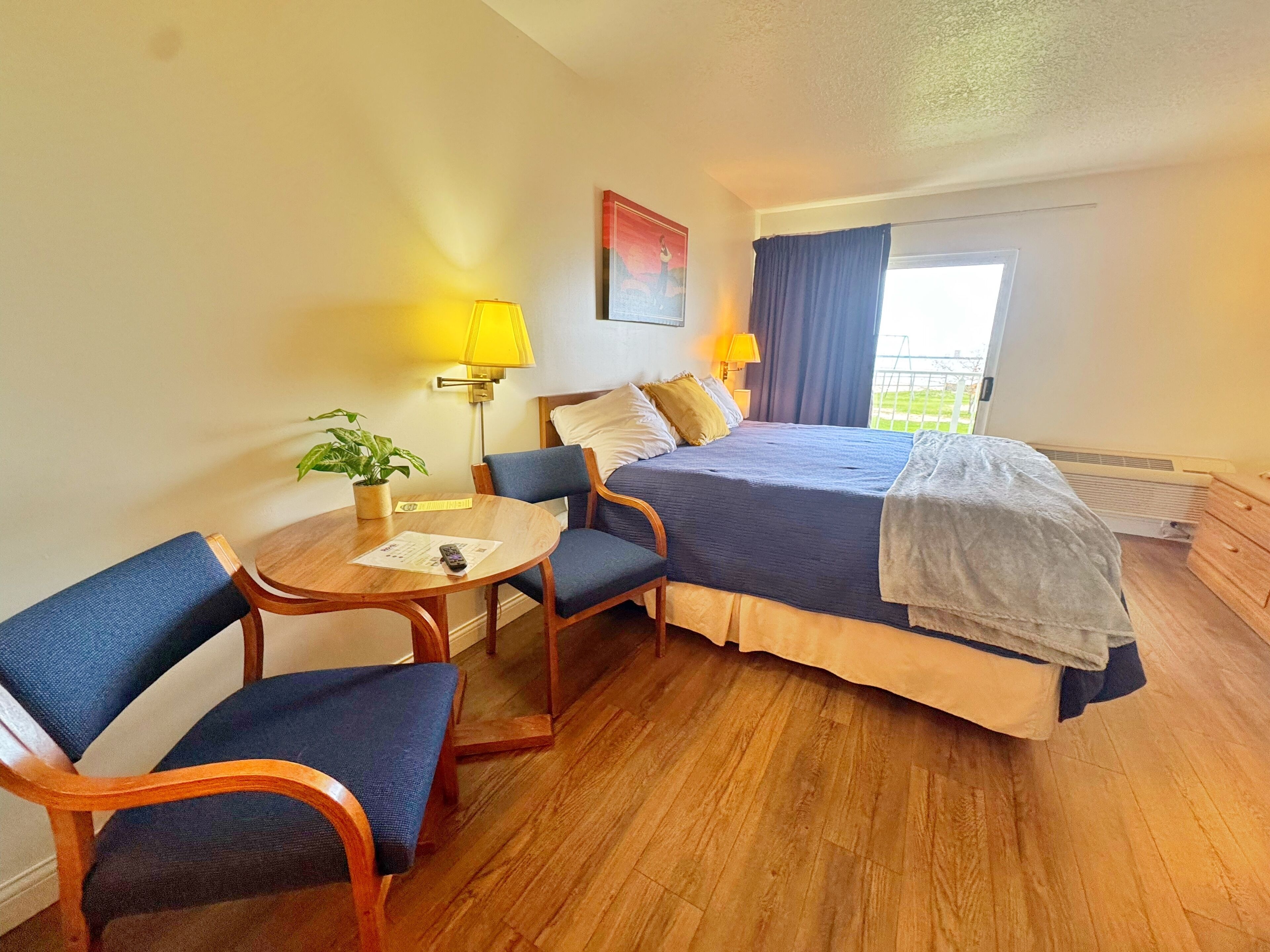 Habitación de hotel con cama, dos sillas, una mesita con lámpara, una planta en maceta y una ventana con cortinas.