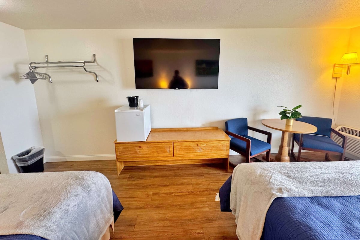 Habitación de hotel con dos camas, un banco de madera, una mesita con una silla, un televisor montado en la pared y un calentador.