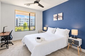 A hotel room with a large bed, two bedside tables with lamps, a desk with a chair, a ceiling fan, and a window with a view of buildings and greenery.