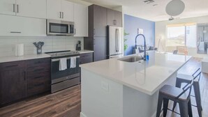 Une cuisine moderne avec des meubles de couleur sombre, un îlot central doté d’un plan de travail blanc, des appareils électroménagers en acier inoxydable et une vue sur l’espace de vie.