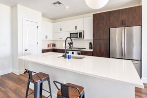 A modern kitchen with white cabinetry, a stainless steel refrigerator, a microwave, and a large island with a sink. There are two bar stools and a coffee machine.