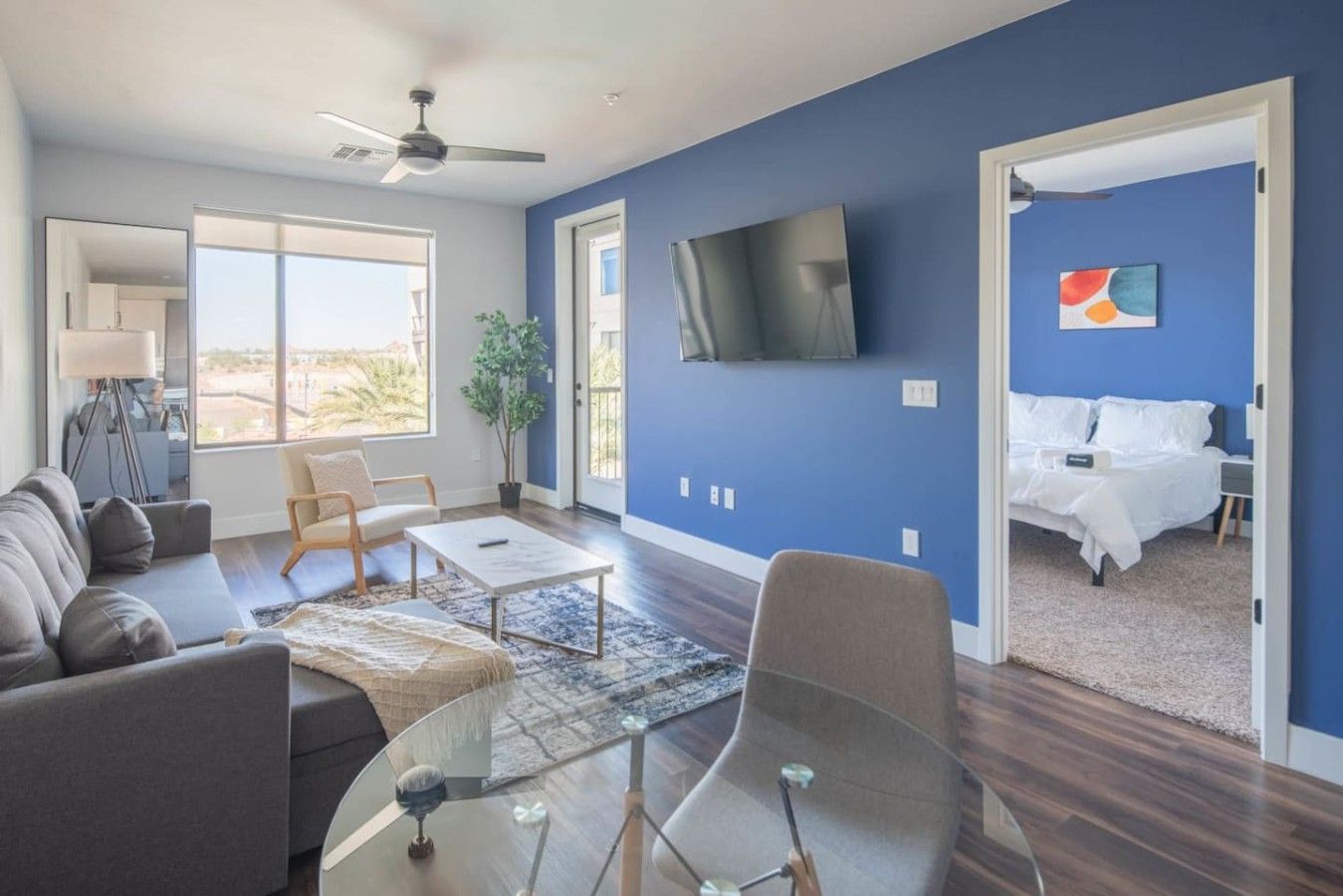 A modern living room with a sofa, coffee table, and TV. There is a bed in the adjacent room with a view.