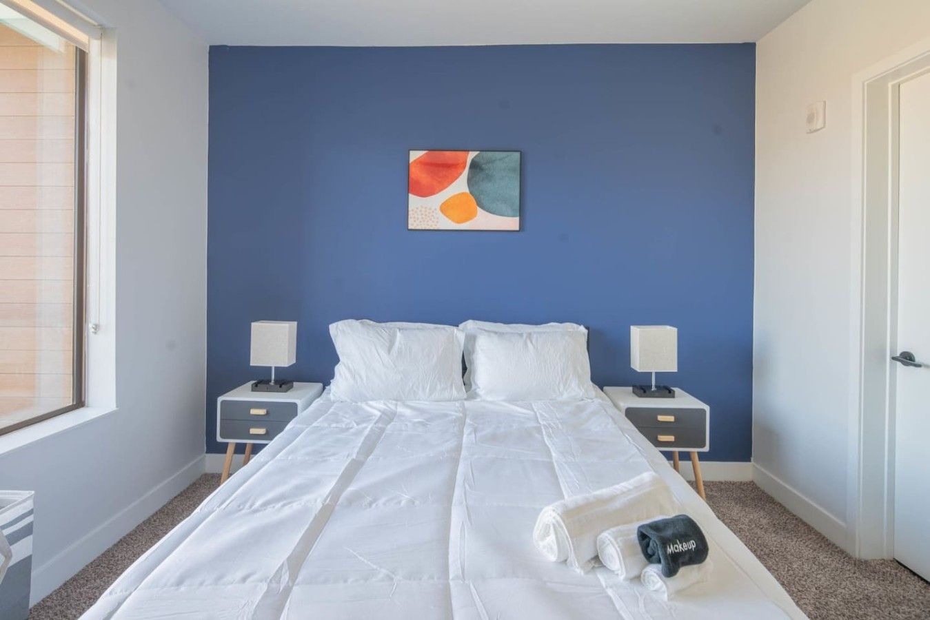 Un dormitorio con una cama grande, dos mesitas de noche, un cuadro abstracto colgado en la pared y una puerta a la derecha.