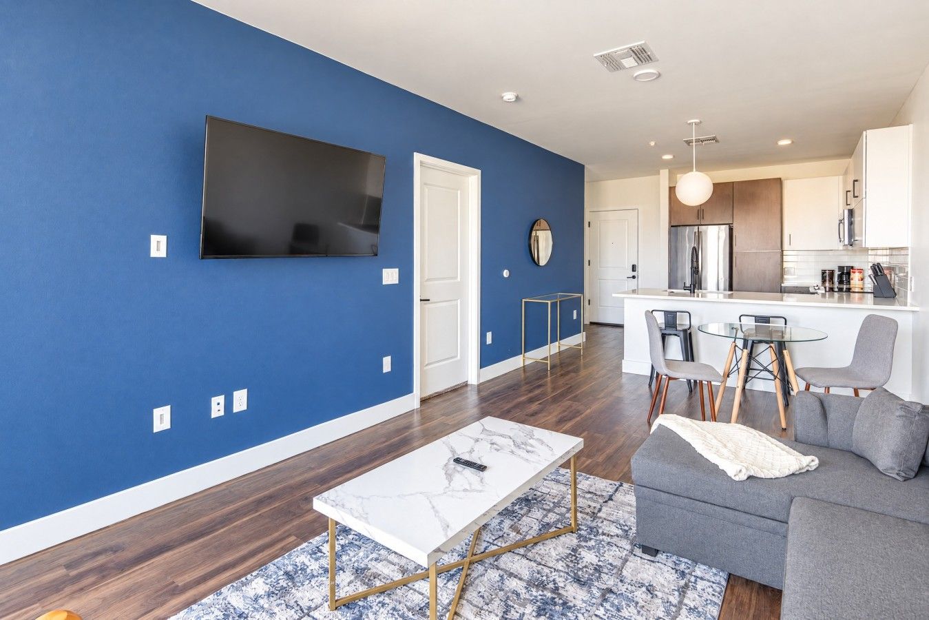 Un salon moderne avec un mur d’accent bleu, un téléviseur à écran plat, une table basse en marbre, un canapé gris et un coin repas avec une table et des chaises en verre.