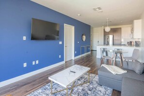 A modern living room with a blue accent wall, a flat-screen TV mounted above a door, a white marble coffee table, and a grey sofa. - Tempe Convenience 1BD with Pool Gym Parking. (Tempe)