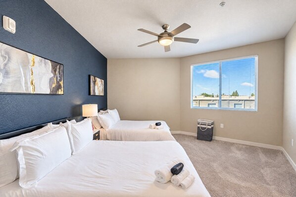 A hotel room with two beds, a ceiling fan, a window with a view, and artwork on the wall.