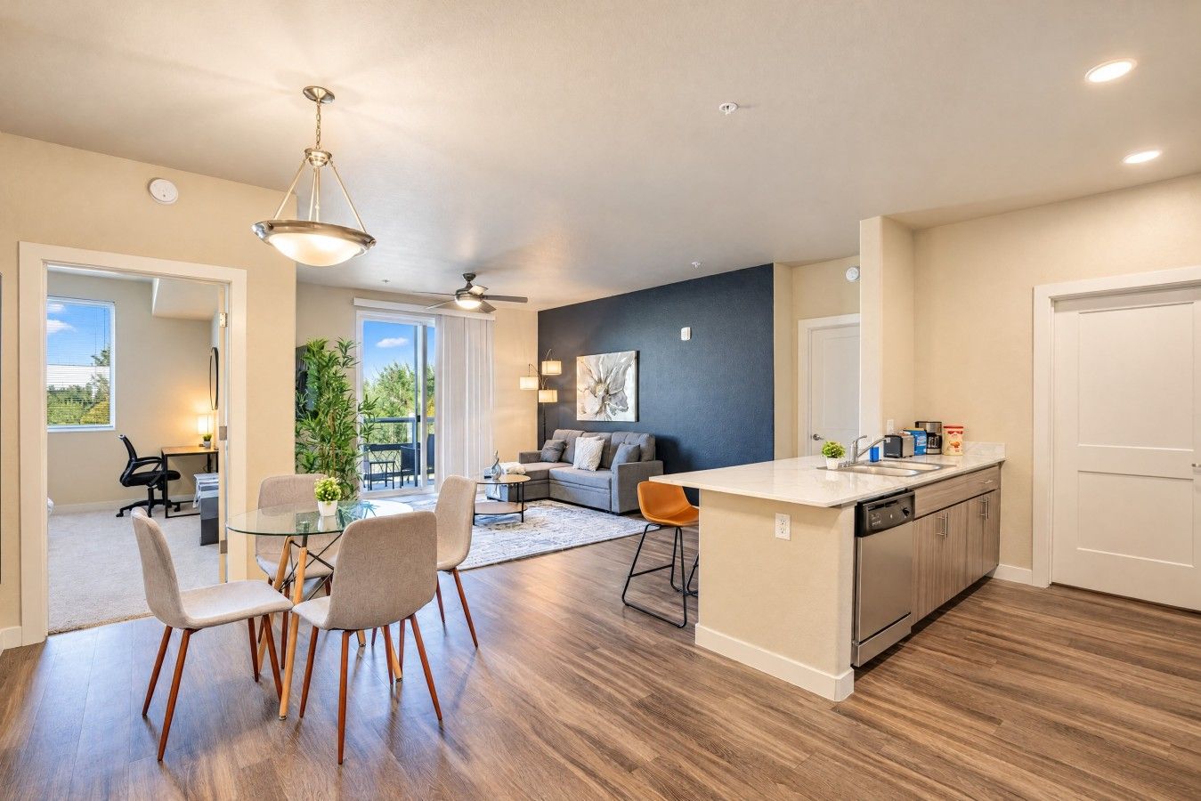 A modern kitchen with a central island, a dining area with chairs, and a living room with a sofa and a ceiling fan.