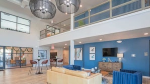 Un hall d’entrée spacieux avec un plafond haut, un coin salon avec un canapé et des fauteuils, un coin repas avec une table et des chaises, et un téléviseur fixé au mur.