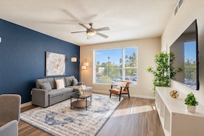A modern living room with a sofa, coffee table, and chairs, a ceiling fan, a large window with a view, and a mounted TV. - DT Tempe Retreat | Pool,Gym,Parking,W/D,Balcony (Tempe)