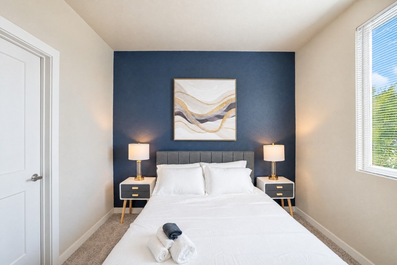 Habitación de hotel con cama, dos mesitas de noche, un aparador, una obra de arte colgada en la pared y una ventana con persianas.
