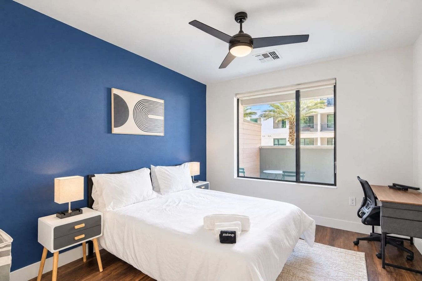 A bedroom with a bed, bedside tables, a desk, a chair, a ceiling fan, and a window with a view of buildings and palm trees.
