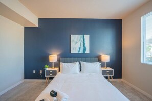 A hotel room with a large bed, two bedside tables with lamps, a dark blue accent wall, and a window with blinds.