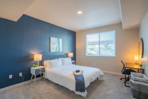 A hotel room with a bed, bedside tables, a desk, a chair, a window with blinds, and a painting on the wall.