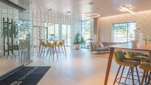 Un hall d’hôtel moderne avec un plafond haut, des cloisons en verre, des chaises jaunes, un canapé et une table basse.