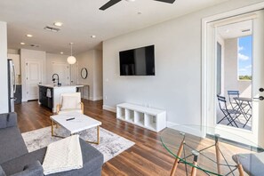 A modern living room with a sofa, coffee table, and a glass table. There is a kitchen area with a dining table and chairs. A TV is mounted on the wall, and a sliding door leads to an outdoor seating area. - DT Tempe 1BD with  Pool Gym Parking WD (Tempe)