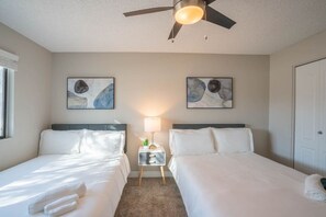 A hotel room with two beds, a ceiling fan, artwork on the walls, a nightstand with a lamp, and a door.