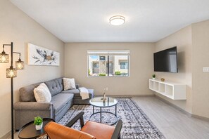 A modern living room with a grey sofa, a coffee table, a TV mounted on the wall, and a large window with a view of the outside.