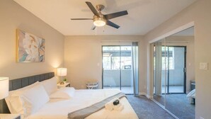 Habitación de hotel con cama, ventilador de techo, ventana y puerta corrediza de vidrio que da a un balcón.