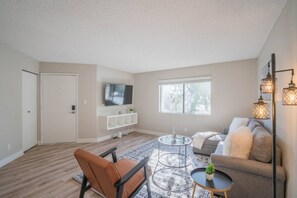 A spacious living room with a sofa, a chair, a coffee table, a TV mounted on the wall, and a window providing natural light. - Walk to Old Town Gym, Pool, WD, Pkg  Balcony (Scottsdale)