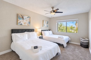 A hotel room with two beds, a ceiling fan, artwork on the walls, and a window with a view of trees.