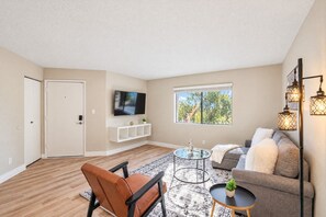 A living room with a sofa, a chair, a coffee table, a TV mounted on the wall, and a window with a view of trees.