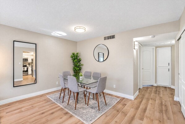 A modern dining area with a glass table, grey chairs, a potted plant, and a mirror on the wall.