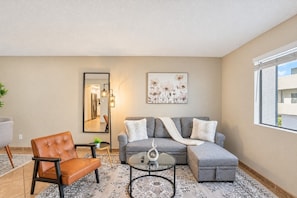 A living room with a grey sofa, a brown leather chair, a round glass coffee table, and a large mirror.