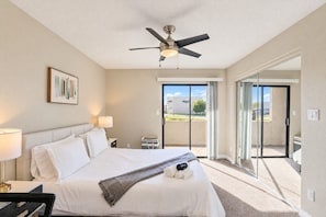 A hotel room with a bed, a ceiling fan, a painting, a nightstand, a lamp, a suitcase, and a view through a sliding glass door.