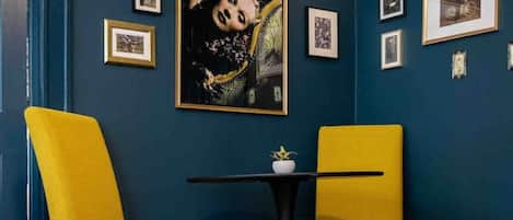 Una habitación con paredes azul oscuro, dos sillas amarillas, una mesita redonda con un jarrón blanco y varias fotografías enmarcadas en la pared.