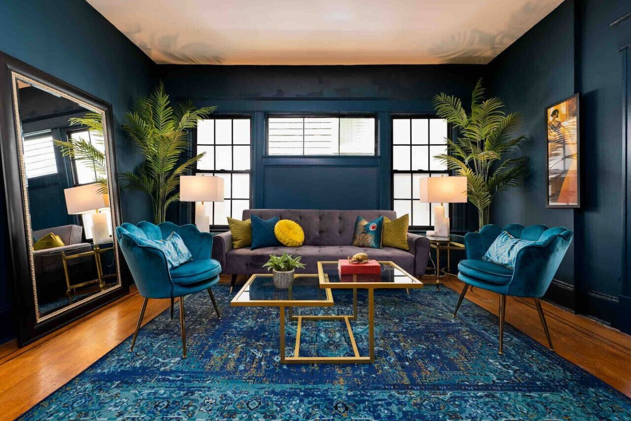A modern living room with a dark blue sofa, two blue armchairs, a glass coffee table, and a patterned rug.