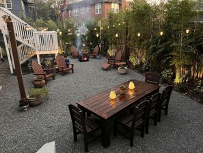 Un patio al aire libre con mesa y sillas de madera, rodeado de plantas en macetas y un brasero.